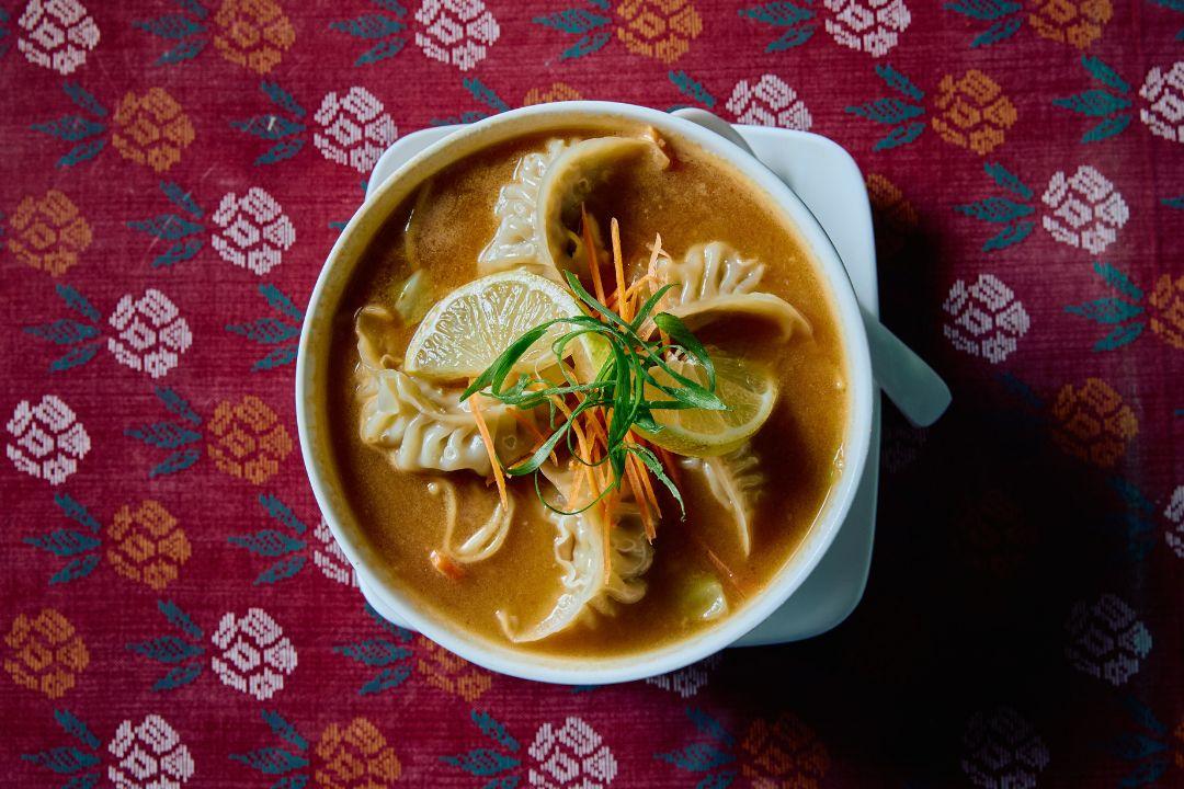 Overhead view of a bowl of thukpa noodle soup topped with steamed dumplings, lime wedges, shredded carrot and sliced spring onions.