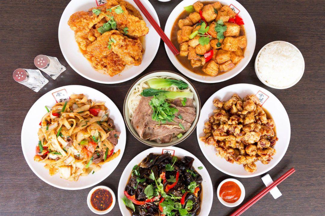 A top-down view of a table featuring various Chinese dishes including stir-fried noodles, crispy chicken, tofu, beef noodle soup, and wood ear salad.