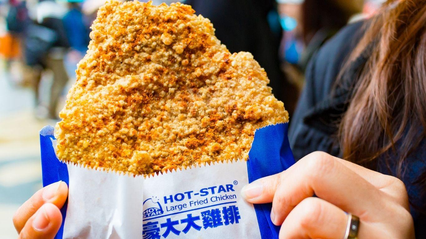 A person holds a large, crispy, breaded fried chicken cutlet in a blue and white paper bag labelled with the Hot-Star Large Fried Chicken logo.