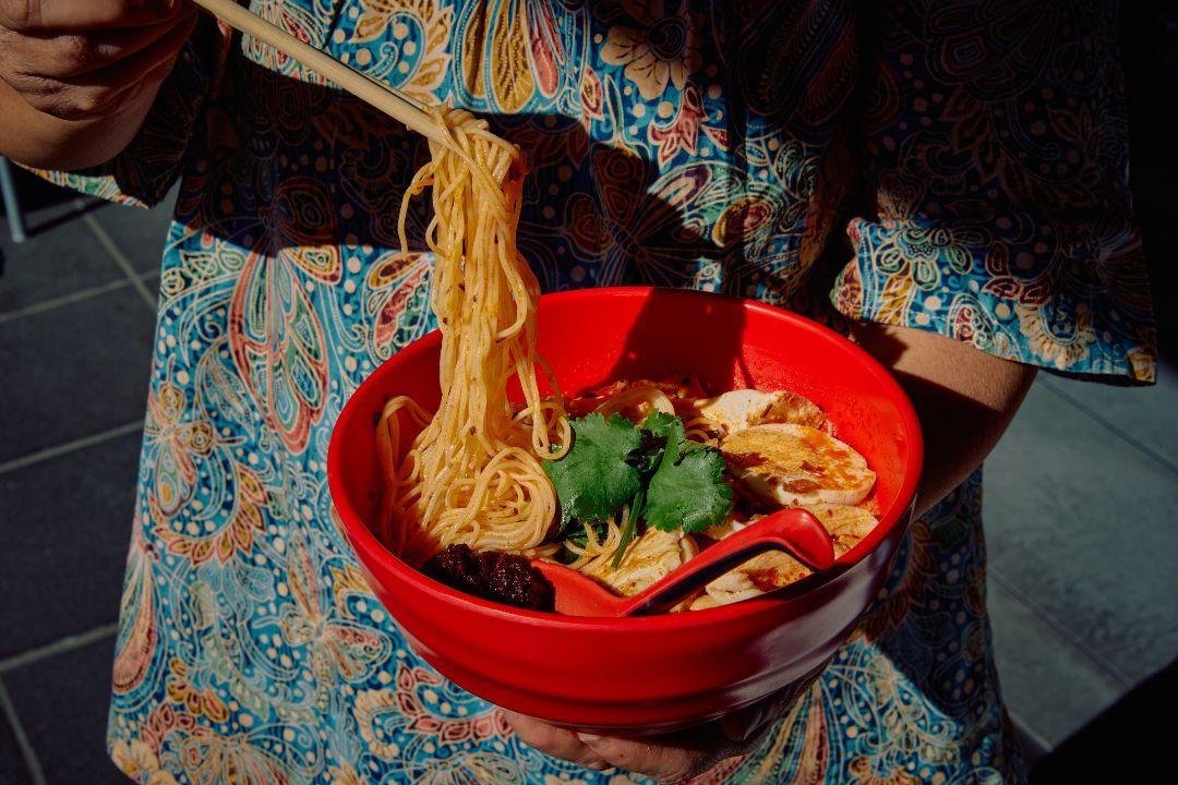 A bright red bowl of laksa is held by a person wearing a colourful patterned shirt.