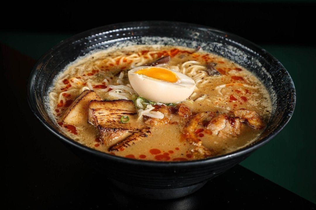 A bowl of ramen in a rich, creamy broth topped with noodles, grilled bamboo shoots, sliced meat and a halved soft-boiled egg.