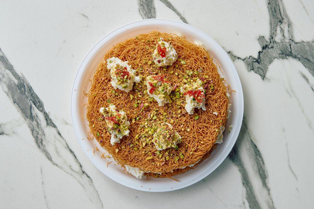 Overhead view of a round Middle Eastern dessert topped with finely shredded golden pastry, dollops of cream and crushed pistachios.
