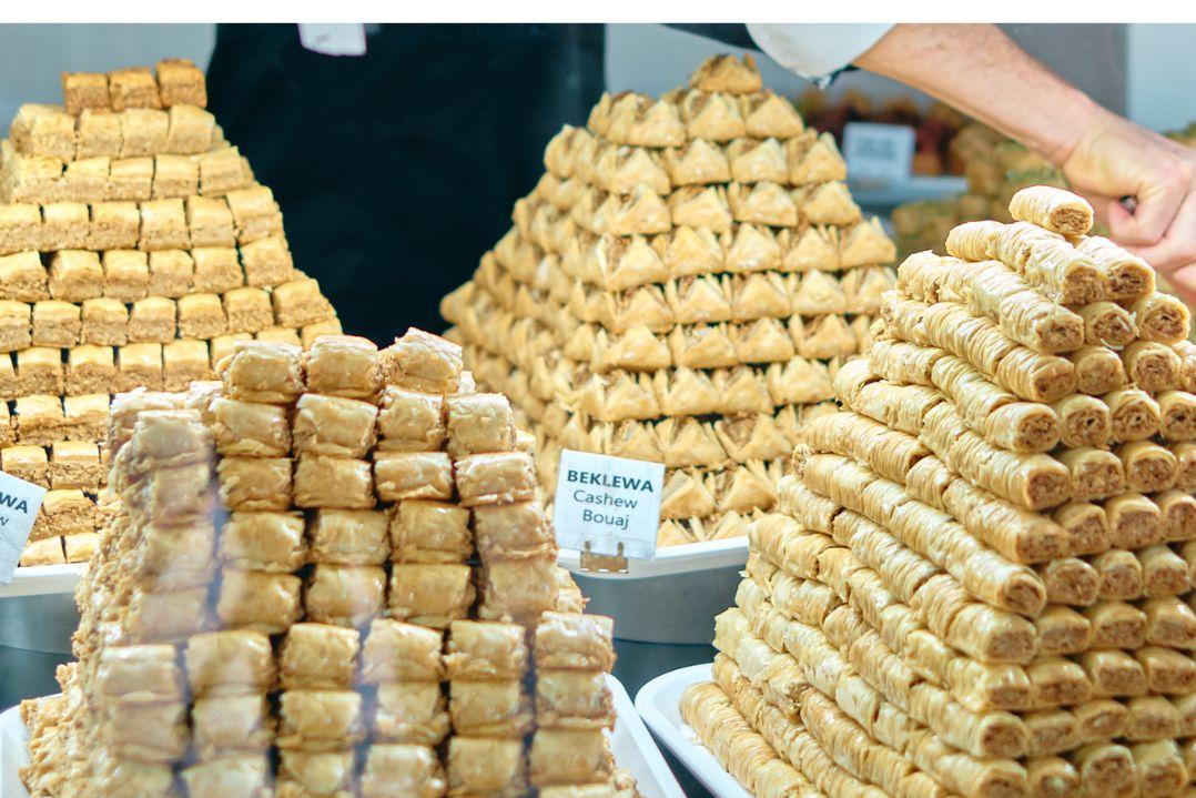 Varieties of baklava, cashew-filled pastries and rolled filo sweets are neatly layered on trays, with small product labels visible among the golden pastries and a baker serving customers in the background
