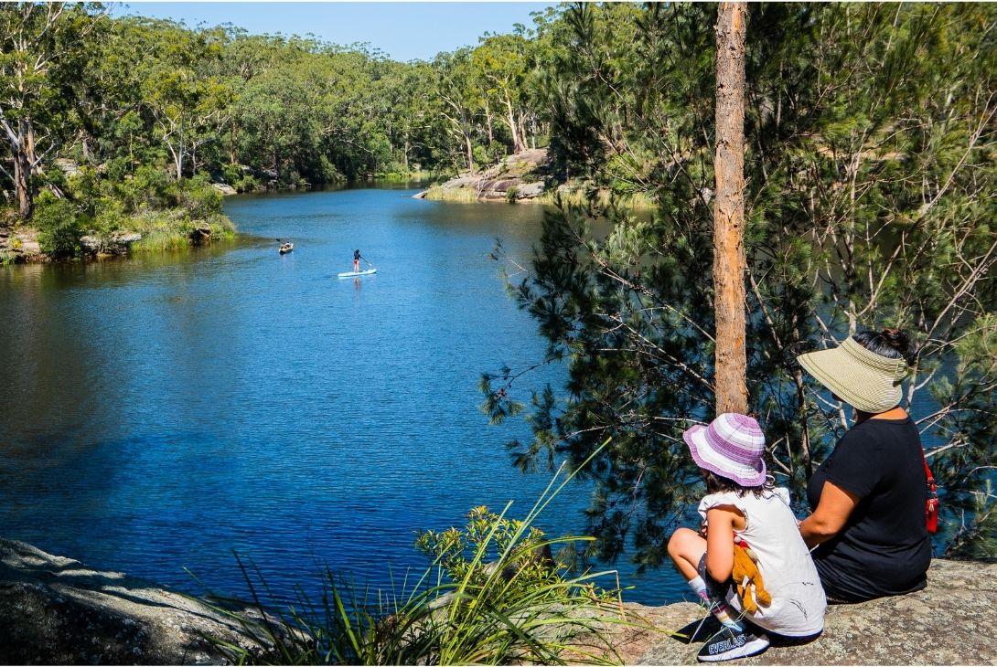 A woman and child sit on a rock, gazing at a flowing river surrounded by lush greenery.