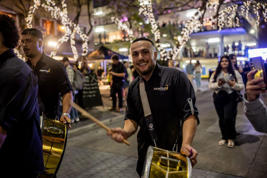 A smiling drummer in a black shirt plays a gold snare drum at an outdoor evening event, with string lights and blurred people in the background.
