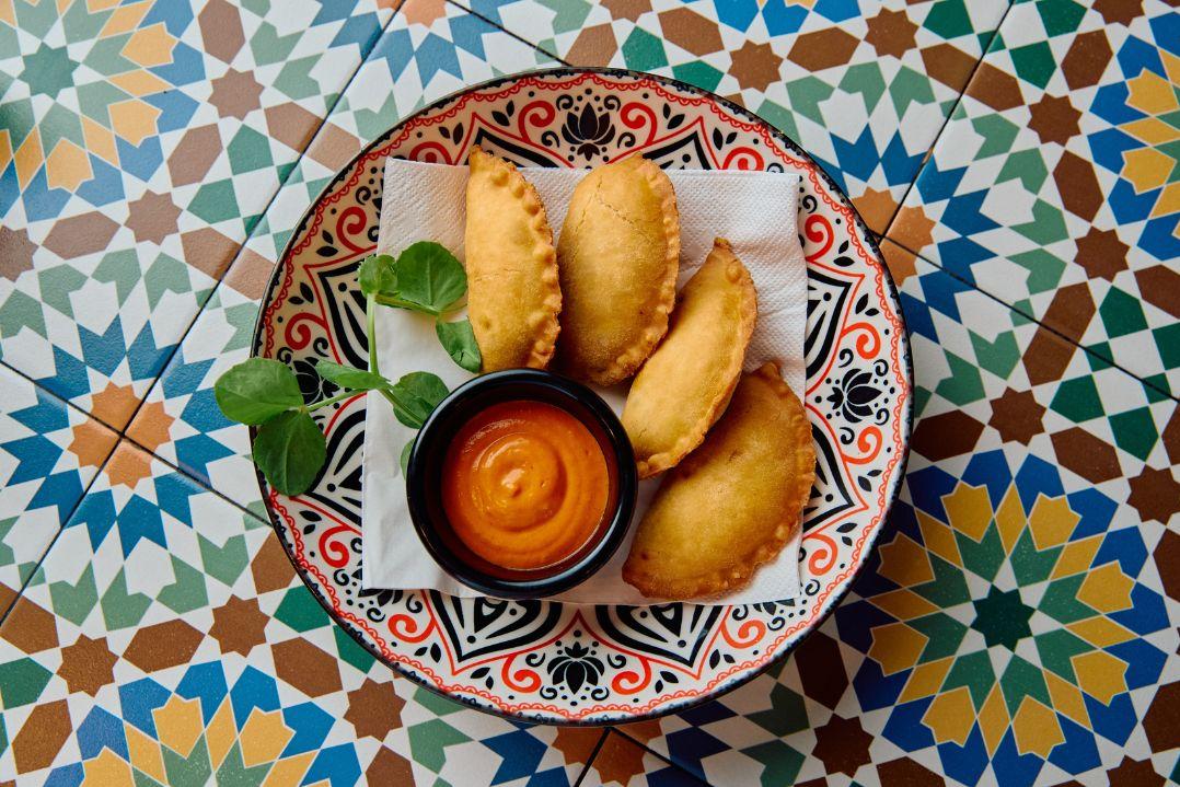 Overhead view of a decorative plate holding five golden fried empanadas served with a small bowl of dipping sauce.