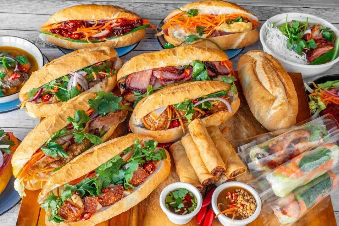 Selection of Vietnamese dishes arranged on a wooden table, including several bánh mì rolls filled with grilled meats, pickled vegetables, fresh herbs and chilli.