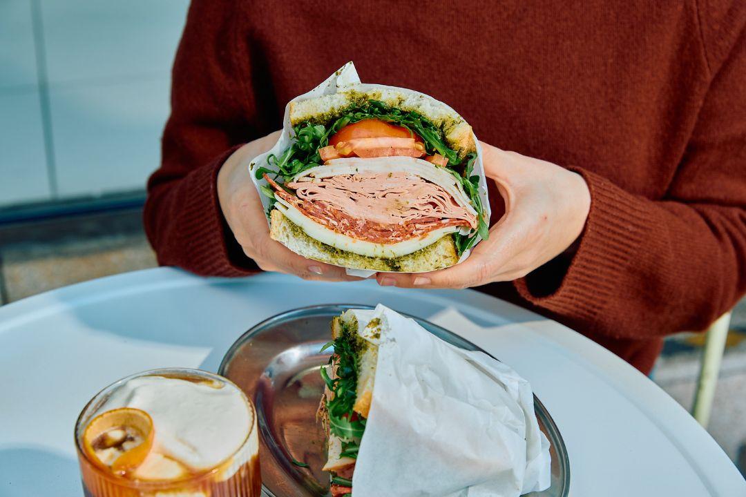 Person holding a large deli-style sandwich filled with sliced meats, cheese, tomato, leafy greens and herb spread while seated at an outdoor café table.