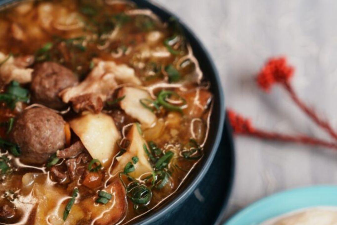 Close-up of a hearty bowl of soup filled with meatballs, mushrooms, vegetables and pieces of meat in a rich broth.