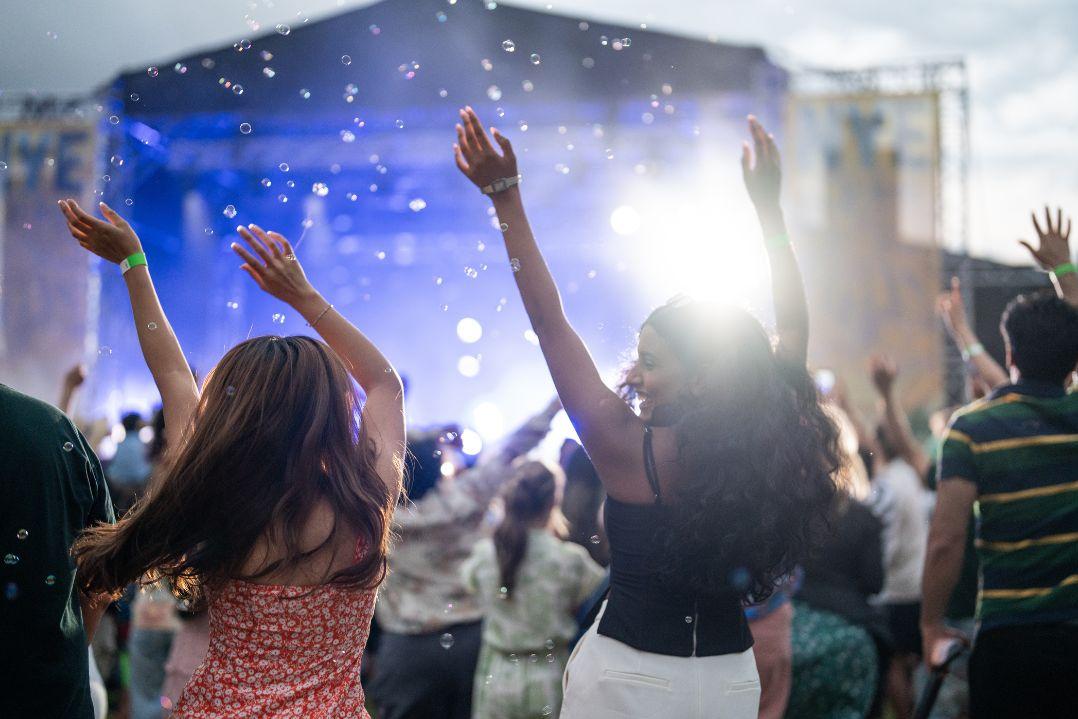 A lively crowd at an outdoor concert, people with hands raised, facing a brightly lit stage under a blue sky, capturing excitement and joy.