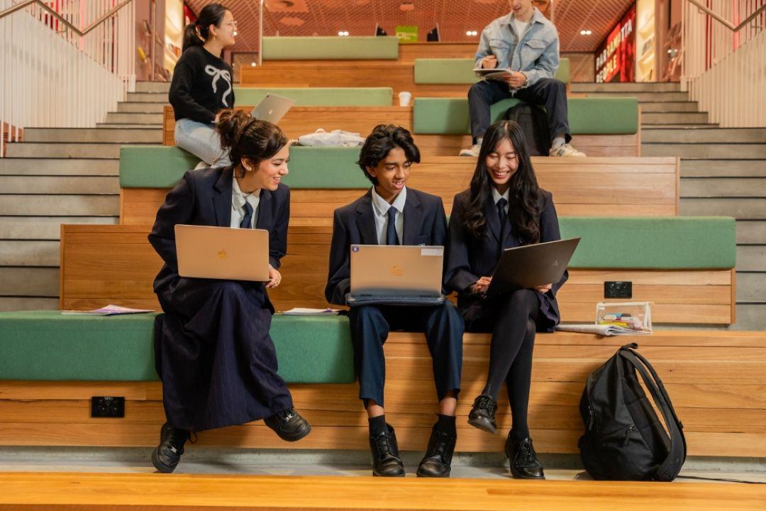 Five diverse students collaborate on laptops in a colorful tiered university lecture hall, creating a friendly, focused atmosphere.