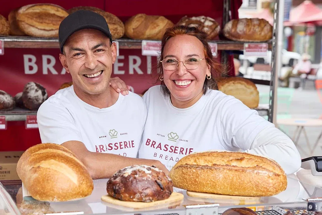 A couple stands in front of a bakery display, showcasing an array of baked goods, including cakes and pastries.