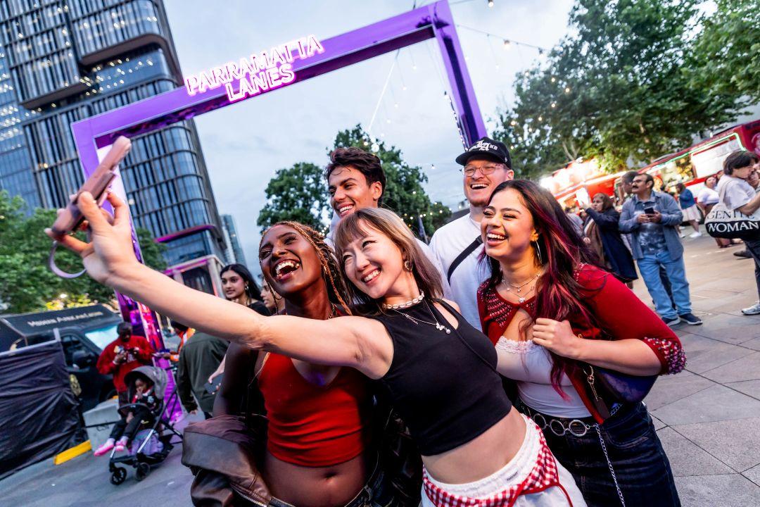 A group of five people happily take a selfie outdoors under a purple frame. They are smiling and the scene exudes a lively, fun atmosphere.