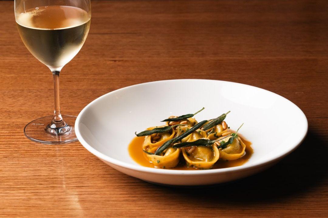 A small serving of tortellini pasta in a light sauce is presented in a large white bowl on a wooden table.