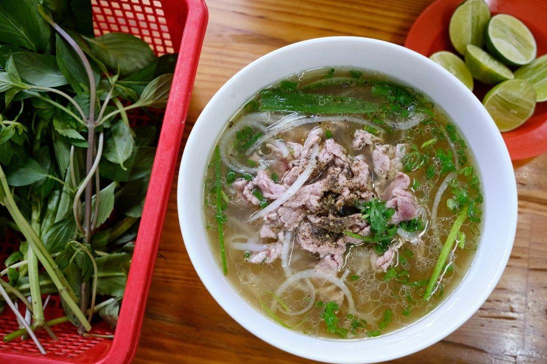 A bowl of beef pho with sliced onions and herbs, served with a side of fresh basil in a red basket and a small plate of lime wedges on a table.