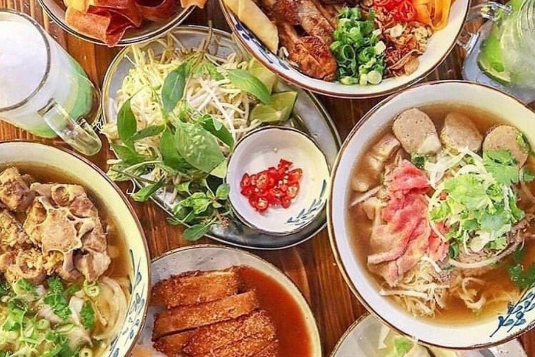 A top-down view of a wooden table filled with bowls of Vietnamese pho, plates of fresh herbs, bean sprouts, sliced chili, and crispy fried meat.