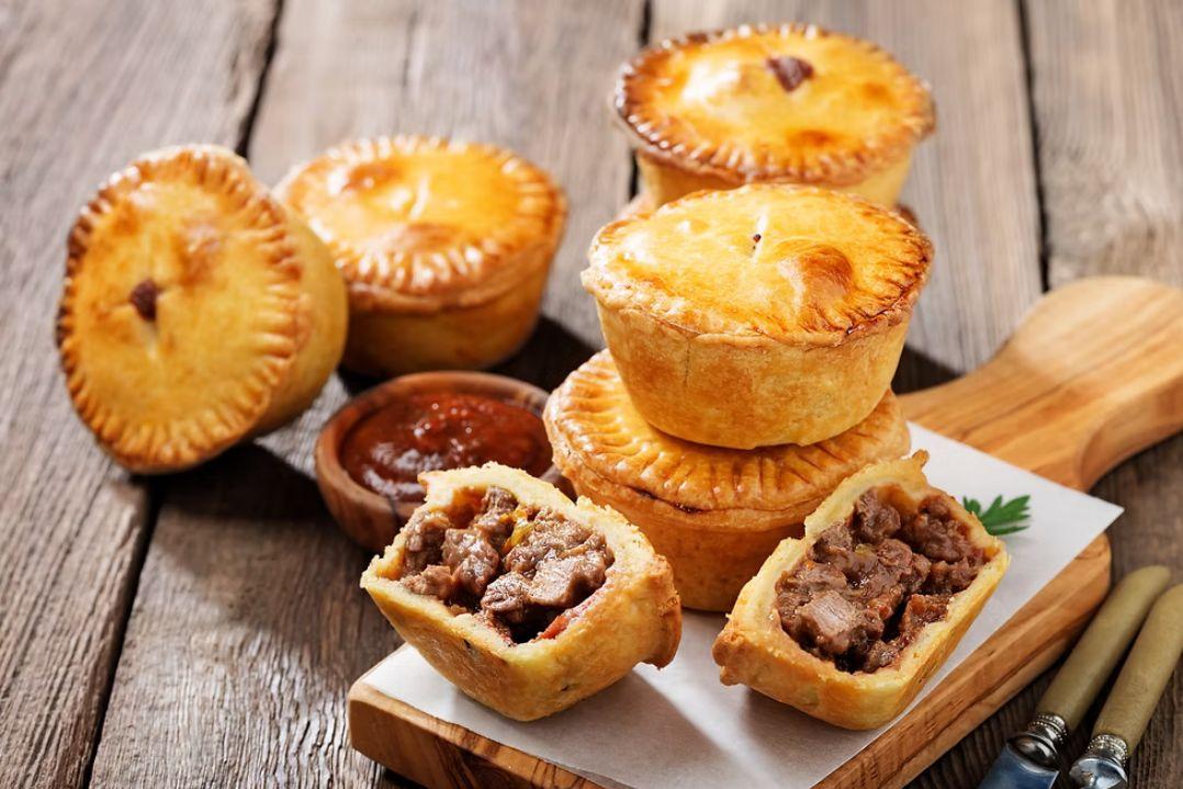 Assortment of golden-brown savoury meat pies arranged on a rustic wooden table. Several pies are stacked on a wooden serving board, while two cut-open pies reveal chunky meat filling.