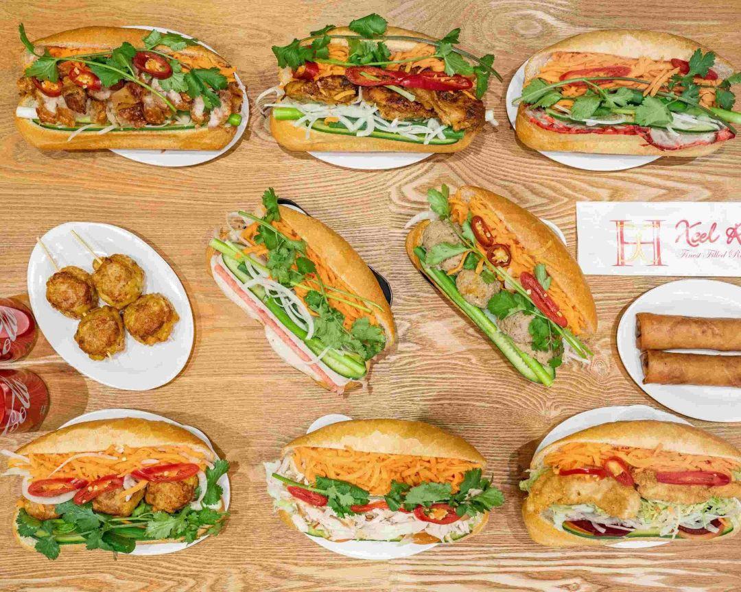 A top-down view of several banh mi sandwiches, a plate of meatballs, and two spring rolls arranged on a light wood table with a branded paper sign.