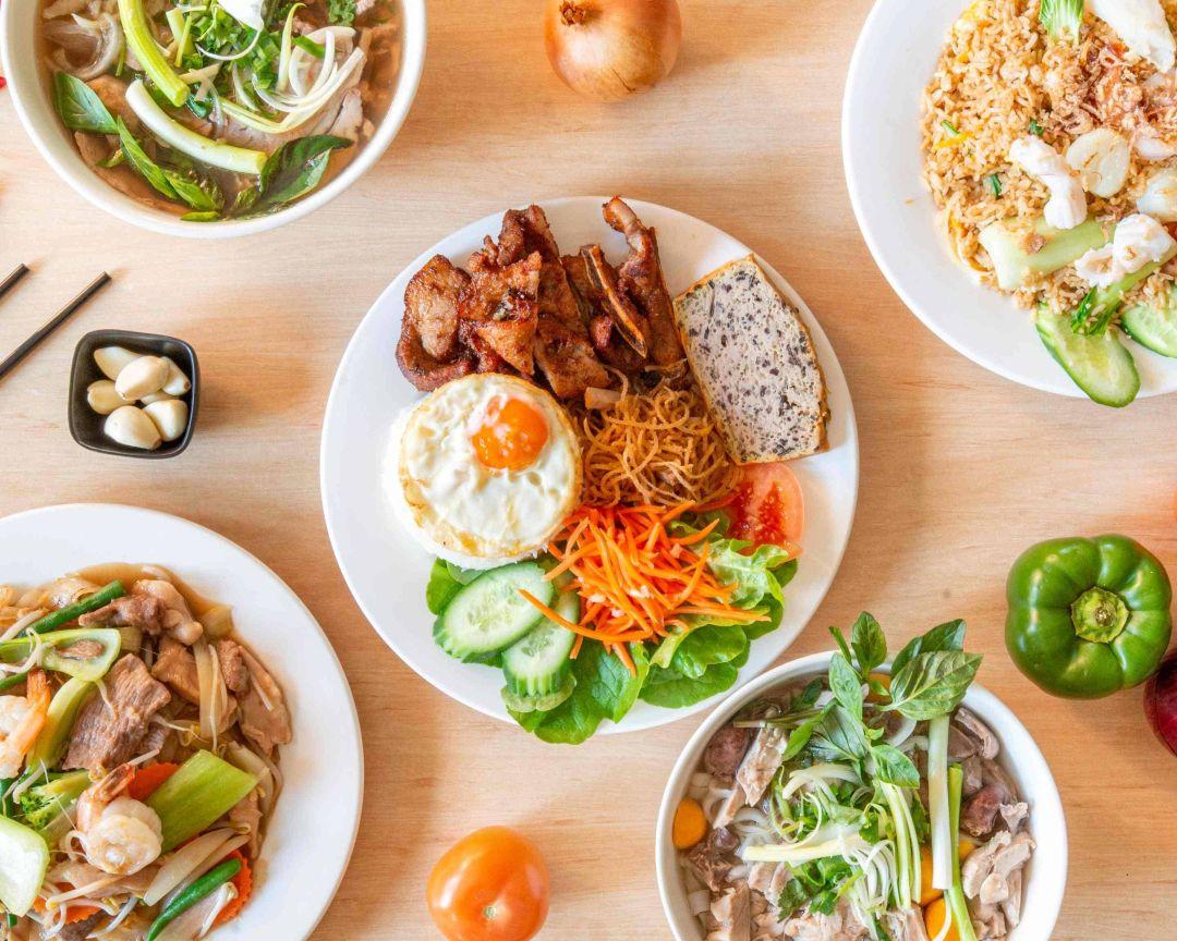 A top-down view of a wooden table featuring a central plate of pork, egg, and rice surrounded by bowls of noodle soup, fried rice, and fresh produce.