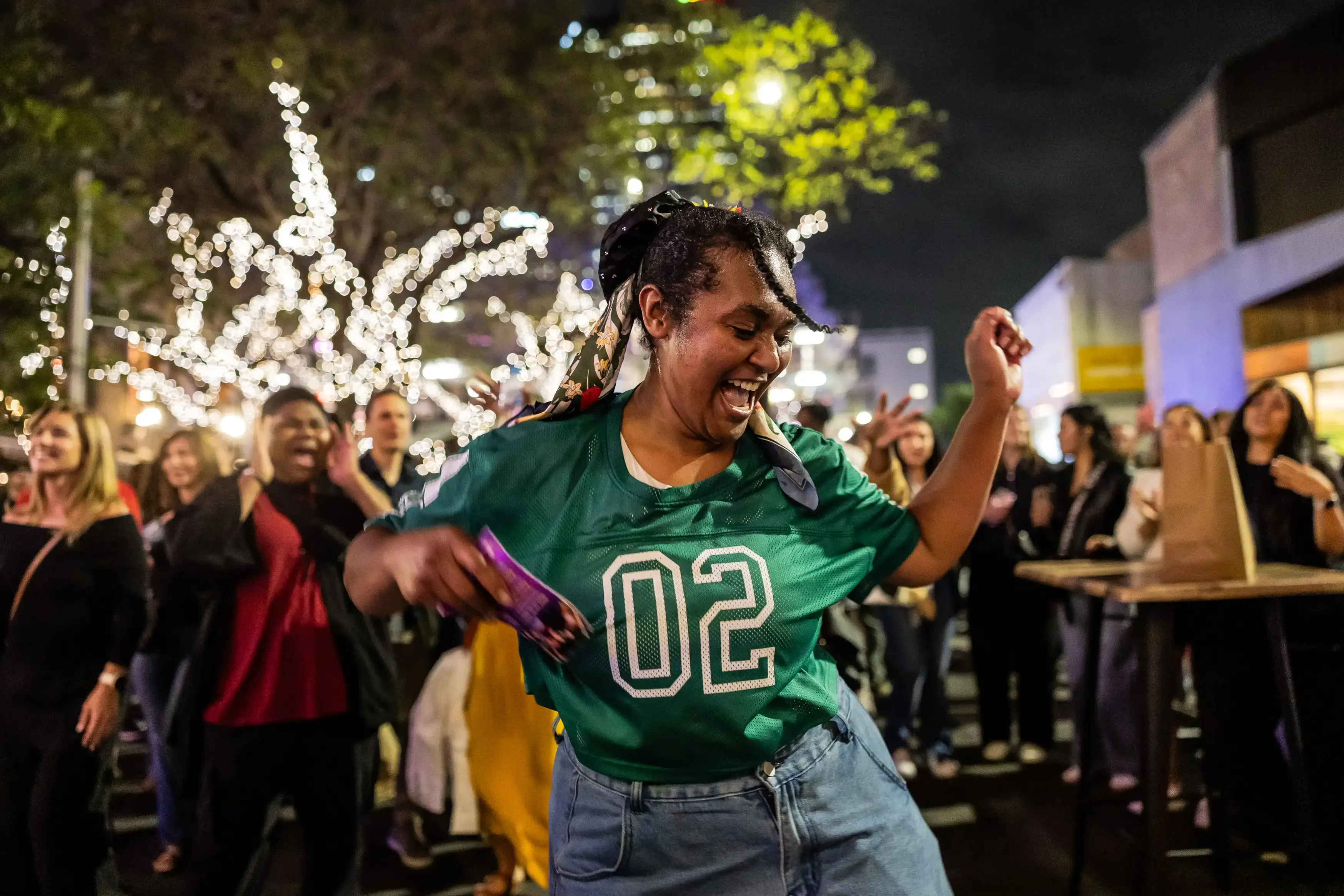 A person in a green jersey dances in the middle of a lively crowd at a nighttime street festival.