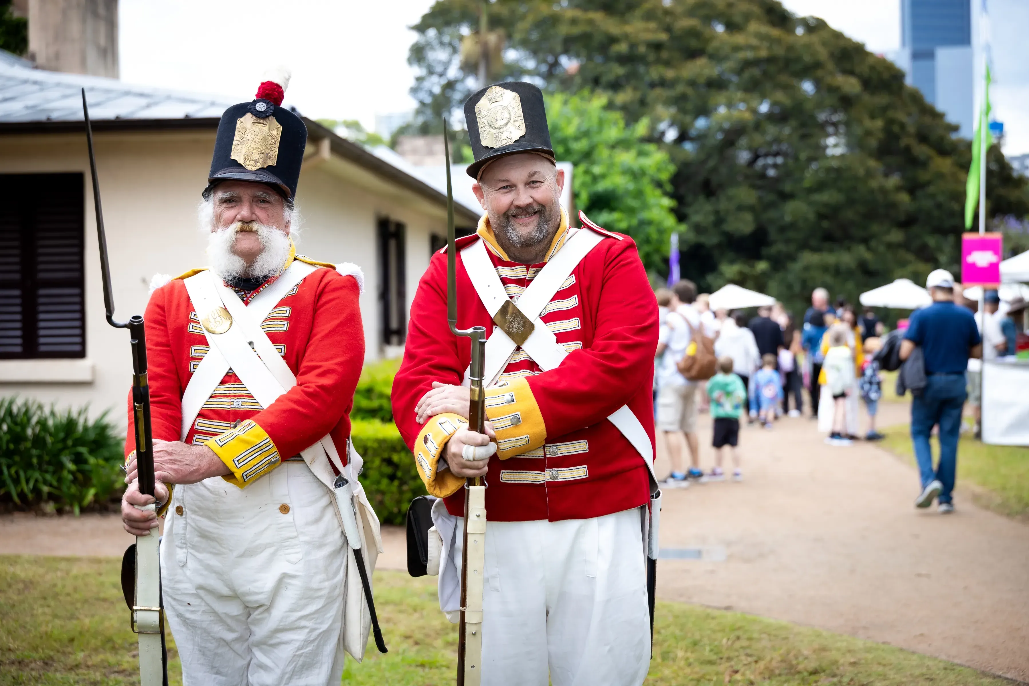 Two historical reenactors dressed in bright red military uniforms and tall black hats stand outdoors holding replica muskets.