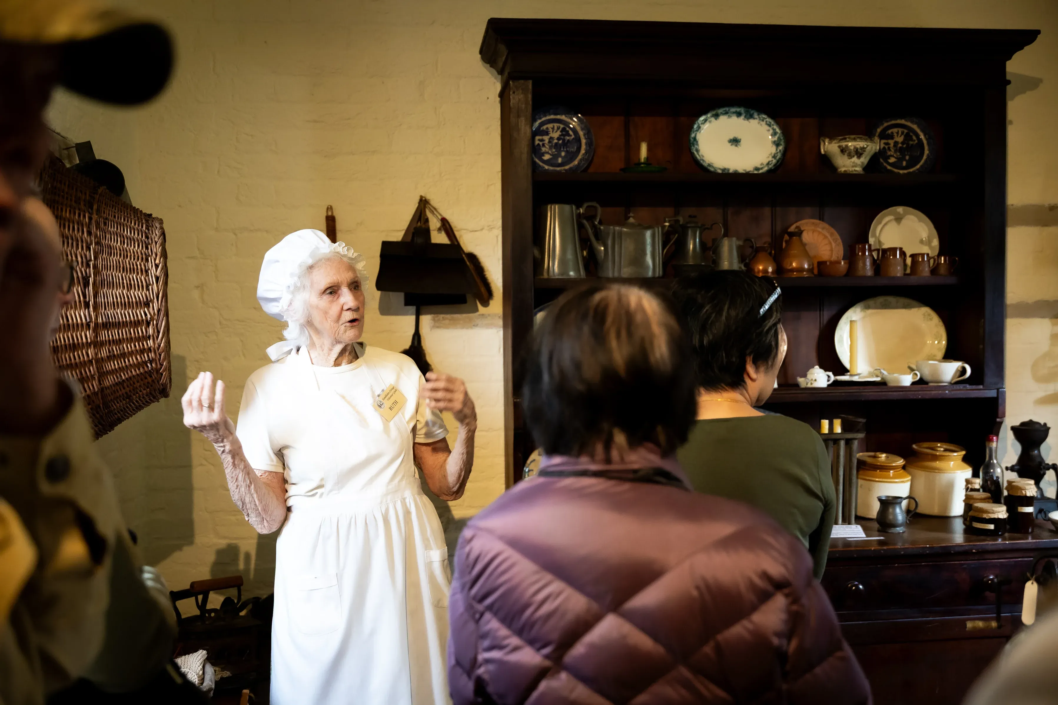 A historical interpreter dressed in a white bonnet and period-style clothing speaks to a group of visitors inside a heritage building.