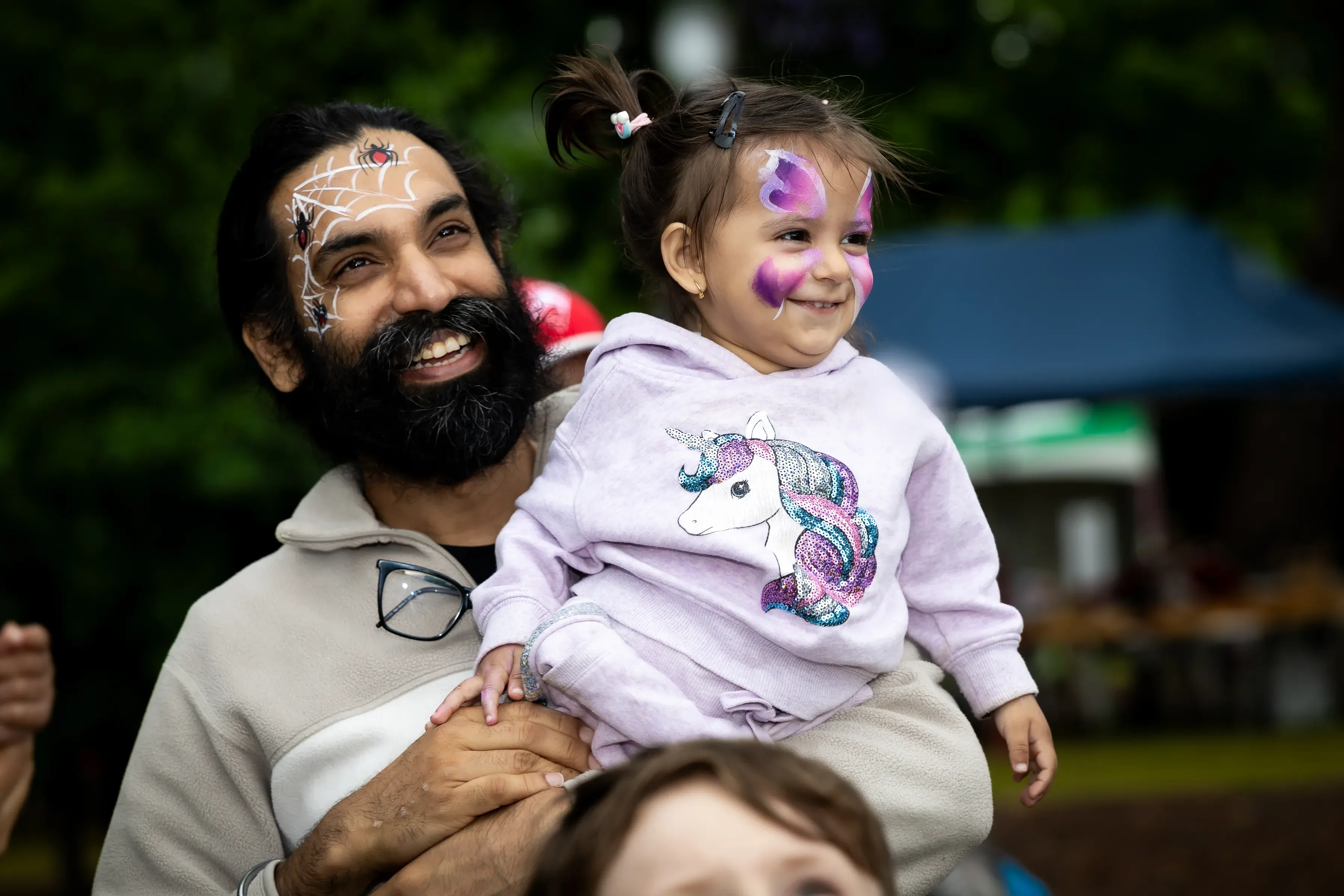 An adult holds a young child at an outdoor community event. Both have colourful face paint, with a spiderweb design on the adult's face and purple butterfly-style face paint on the child's face.