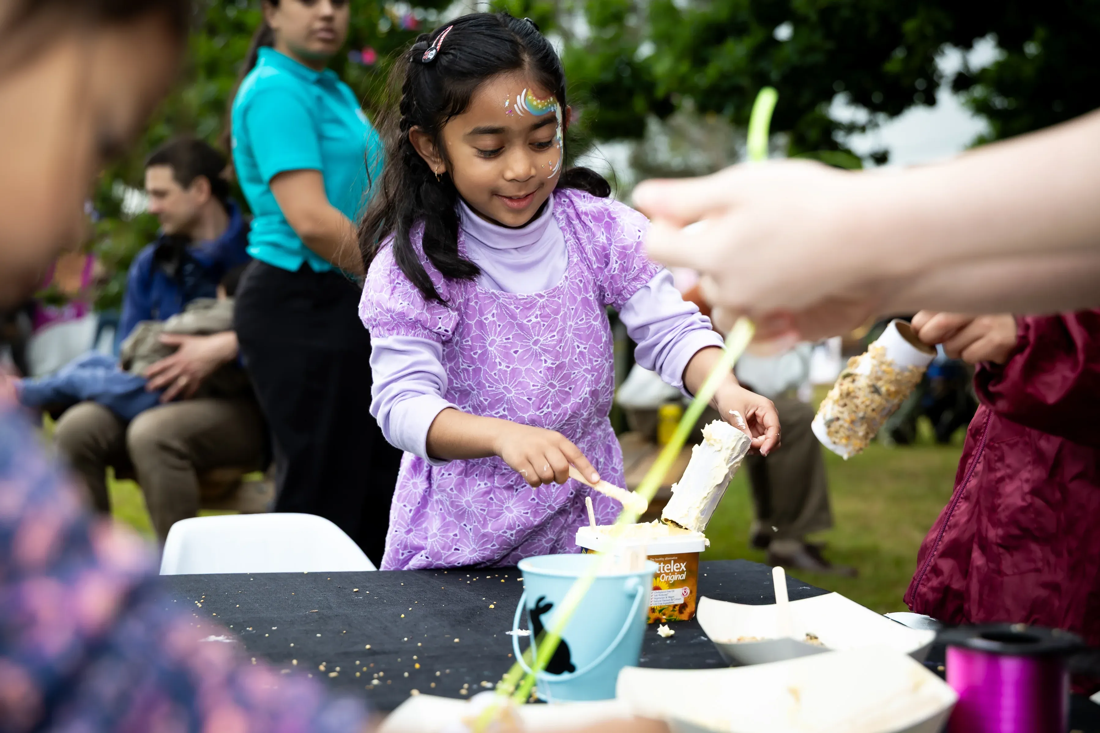 A child with colourful face paint decorates a food item at a community activity table, spreading a topping with a small spatula.