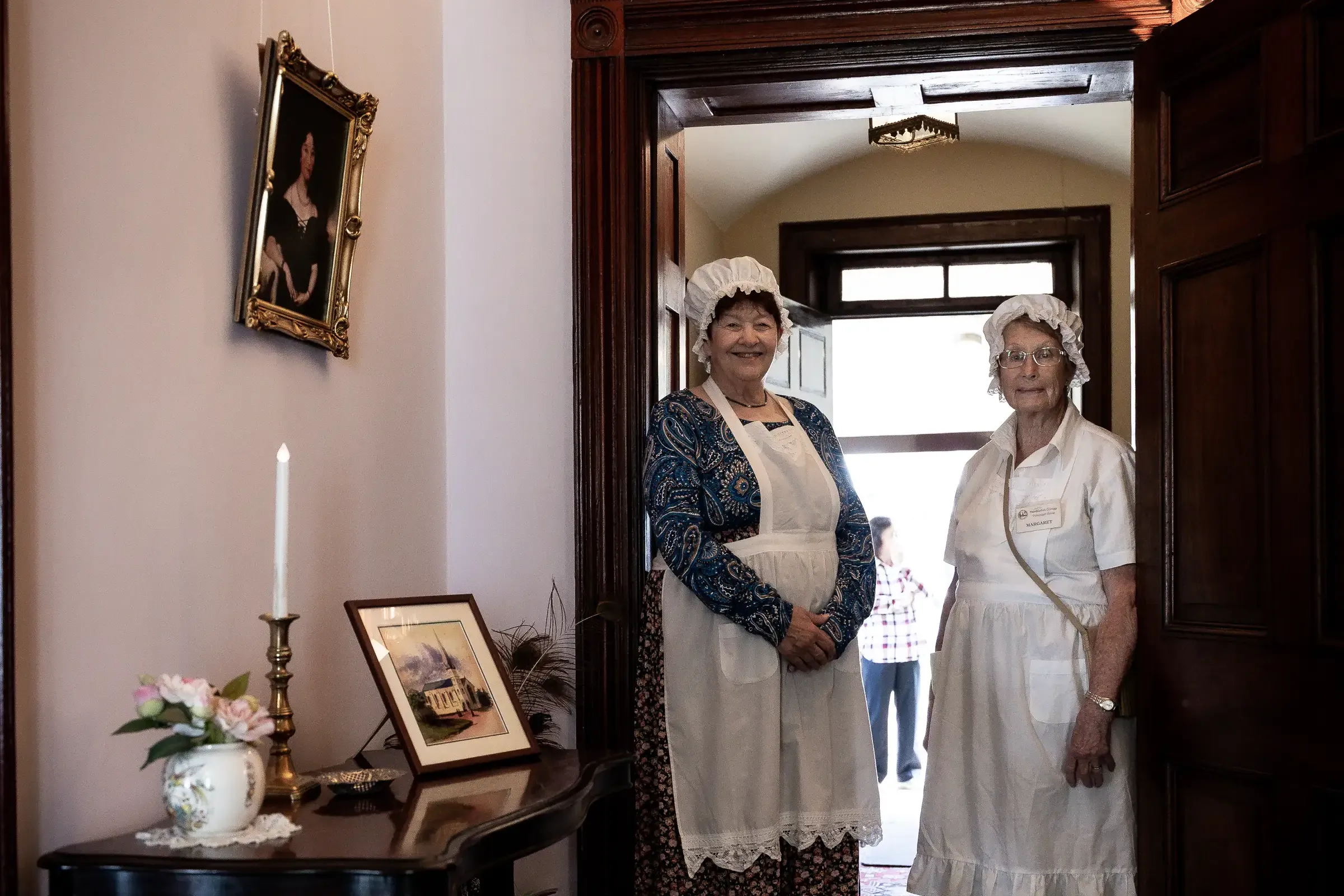 Two volunteers dressed in period-style bonnets, aprons and long skirts stand in the doorway of a heritage house.