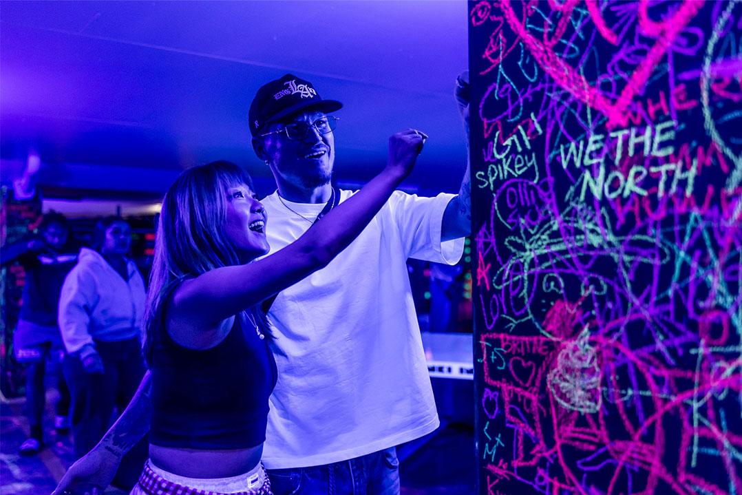 Smiling pair write on a colorful graffiti wall at a blue-purple neon event indoors, surrounded by guests; playful, energetic atmosphere.