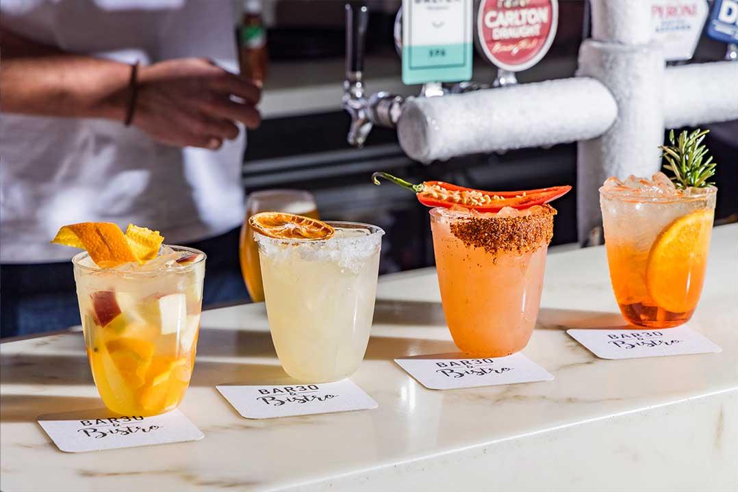 Four cocktails line a bar: fruit-filled yellow, pale, orange, and amber drinks garnished with chips, chili, citrus, and herbs; bartender behind.
