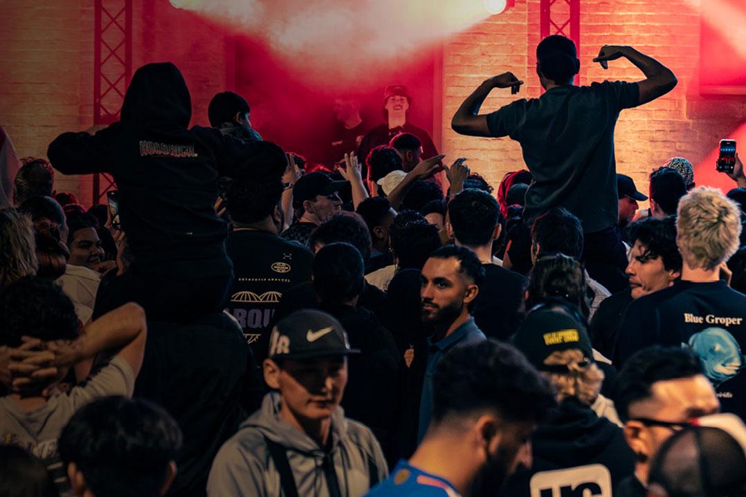 Crowded concert audience in red lighting and smoke, with raised hands, two people lifted above the crowd, and a man flexing.