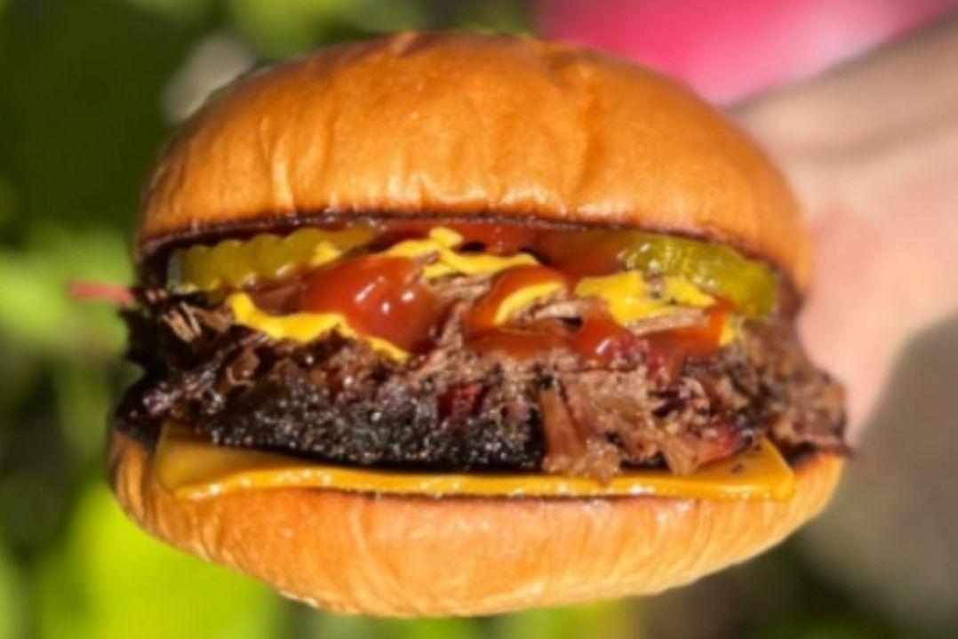 Close-up of a barbecue-style cheeseburger on a toasted bun, filled with smoked pulled meat, melted cheese, pickles, mustard and barbecue sauce, against a blurred outdoor background.