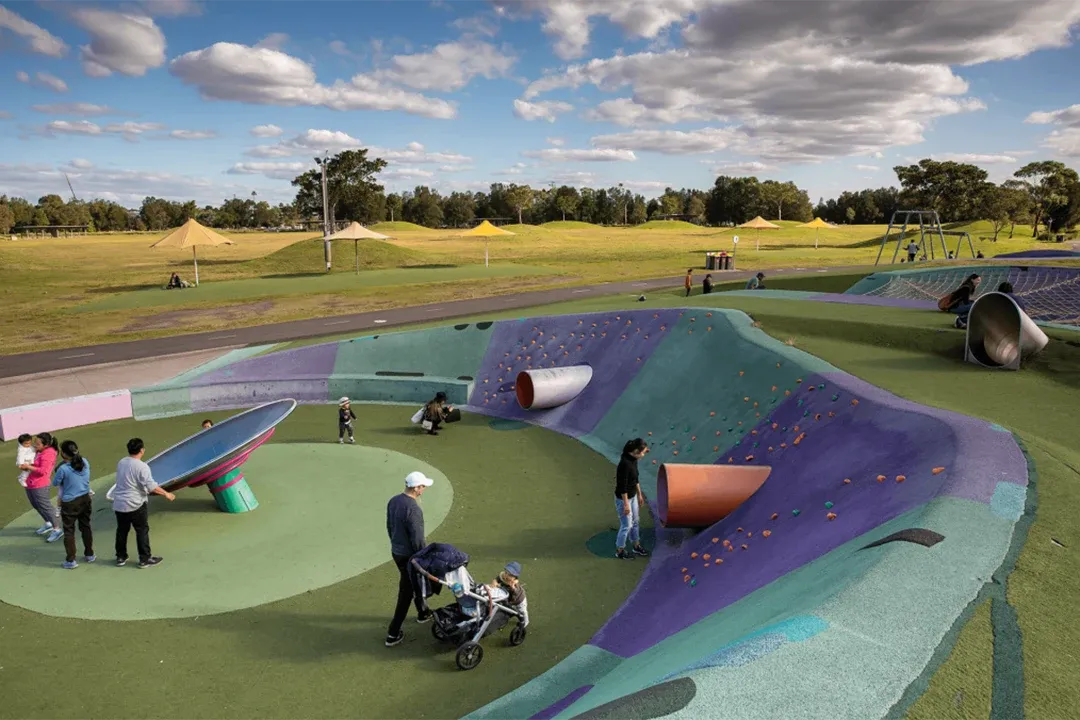 Colourful outdoor skate and climbing park with children and adults using ramps, slides, and play structures under a bright, partly cloudy sky.