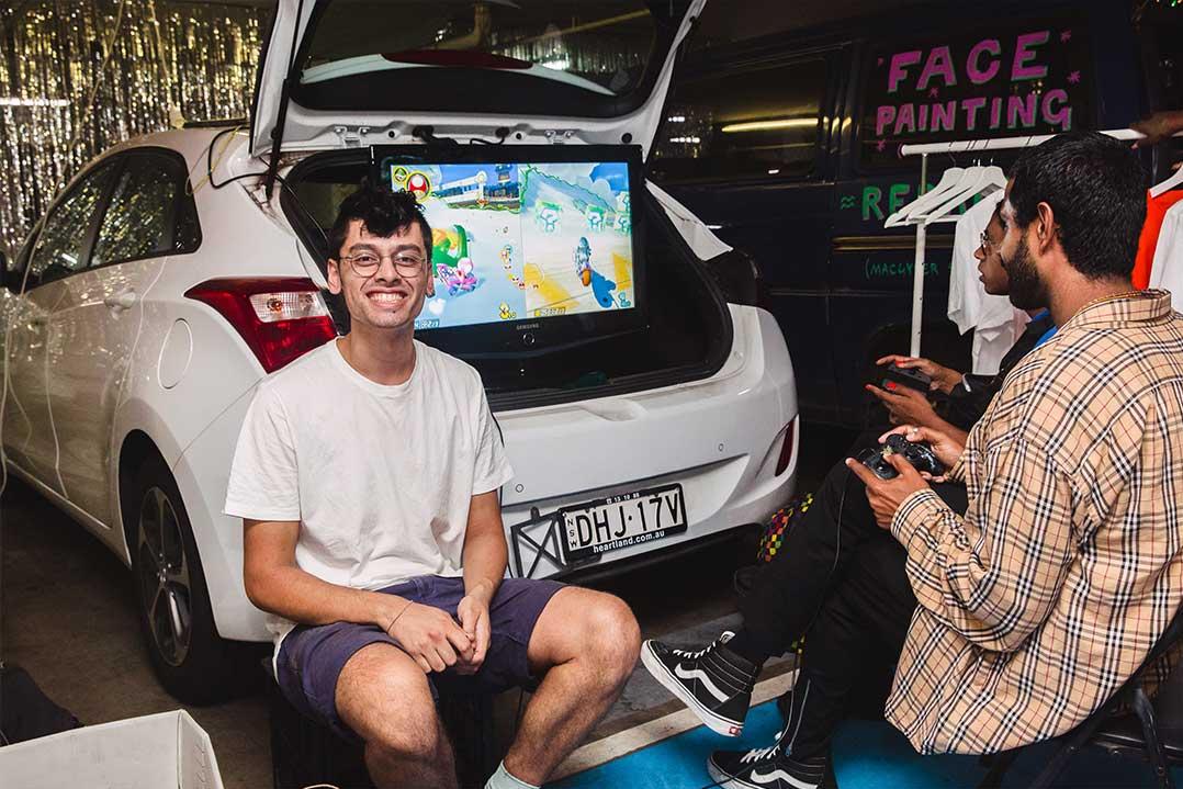 Smiling man sits in an open car trunk beside a gaming screen while another man films him in a colorful, lively auto paint shop.