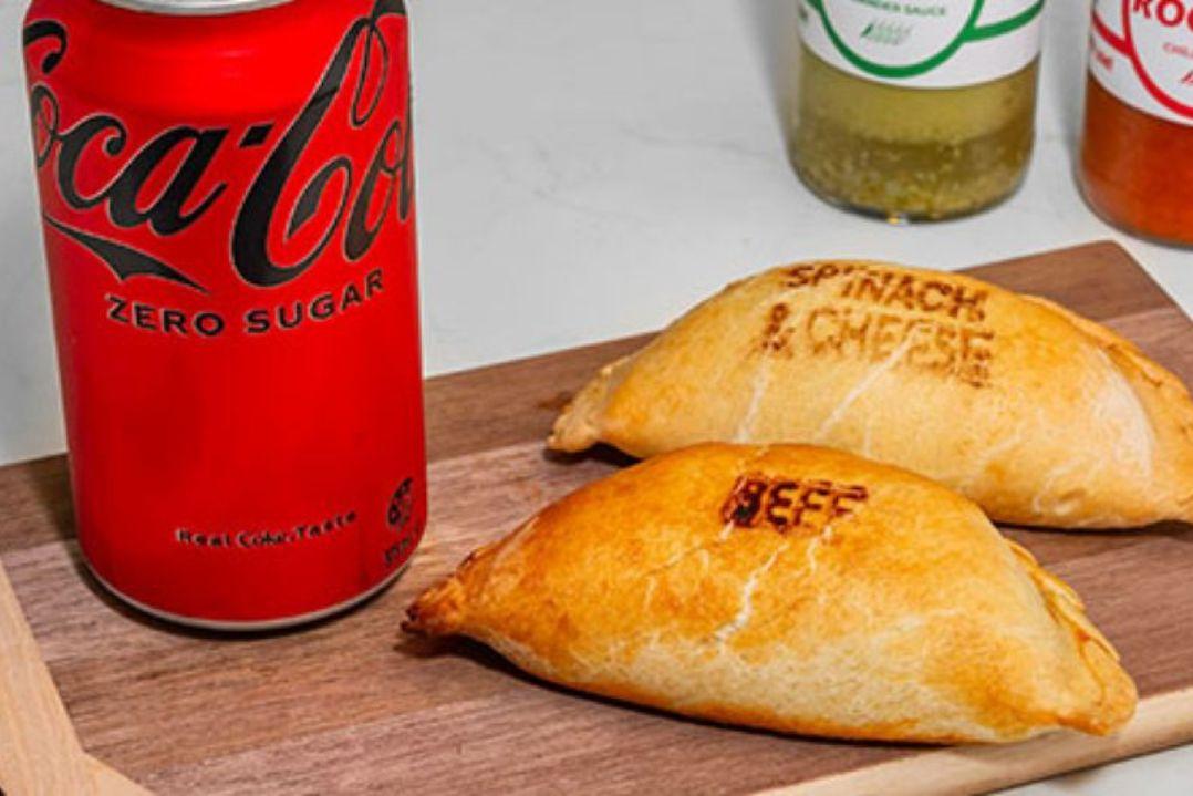 Two baked empanadas labelled “Beef” and “Spinach & Cheese” sit on a wooden serving board beside a can of Coca-Cola Zero Sugar.