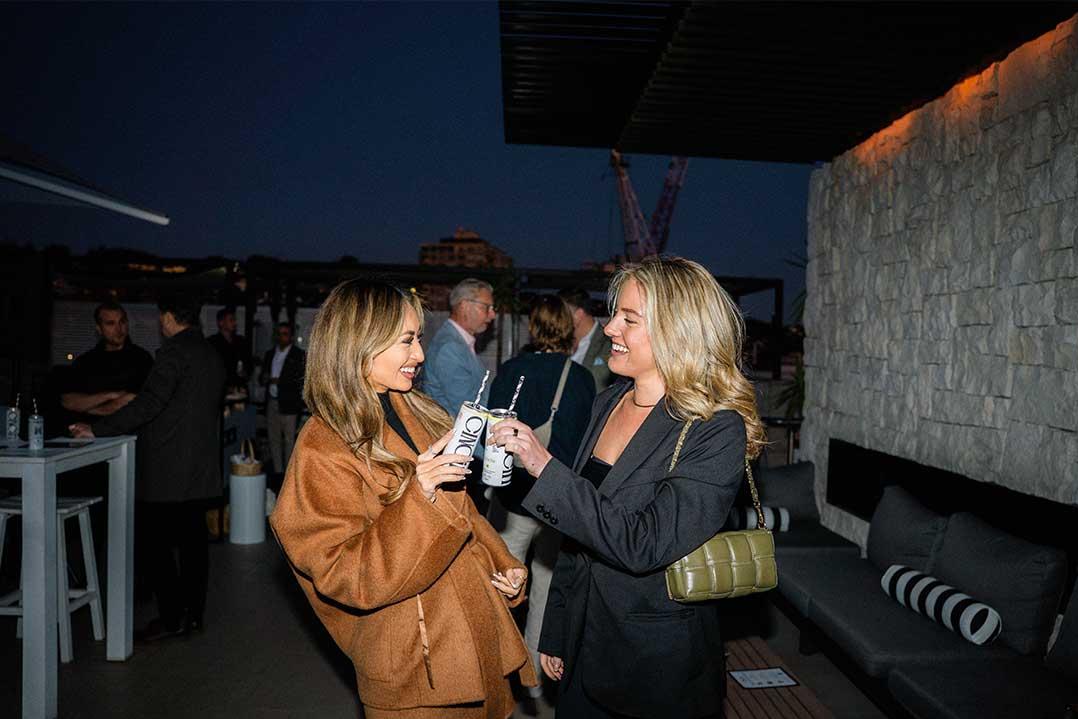 Two smiling women chat at a nighttime rooftop event, one holding a microphone, with guests, drinks, and a stone wall behind them.