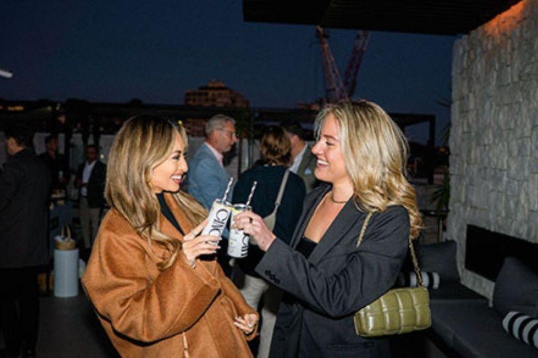Two people standing at an evening social event toast canned drinks while chatting.