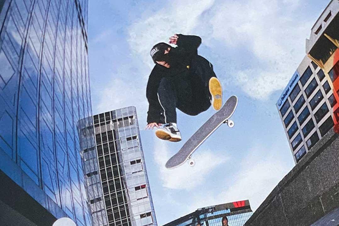 Skateboarder in black clothing soars above a skateboard in an urban plaza, framed by glass office towers under a bright, energetic sky.