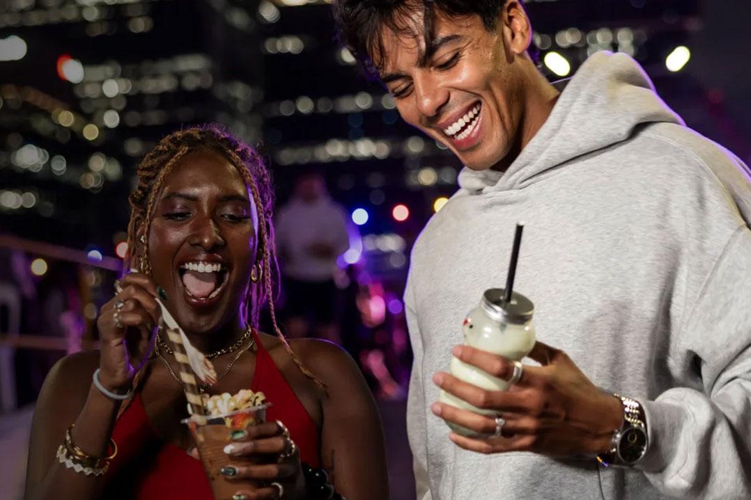 Two friends laugh at a lively nighttime event, holding drinks as colorful city lights and a blurred crowd glow behind them.