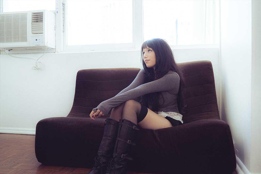 Young woman in dark clothing and knee-high boots sits on a brown sofa in a bright room, gazing thoughtfully toward the window.