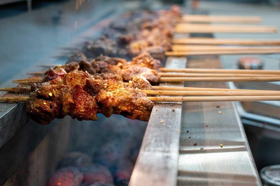 Close-up of spiced meat skewers grilling over glowing charcoal, with smoky heat rising and rows of wooden sticks extending into the background.