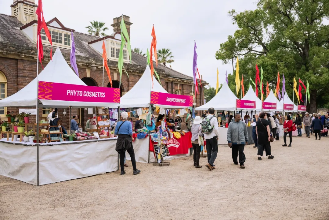 A row of market stalls lines a heritage precinct during a community event, with colourful flags flying above white marquees.