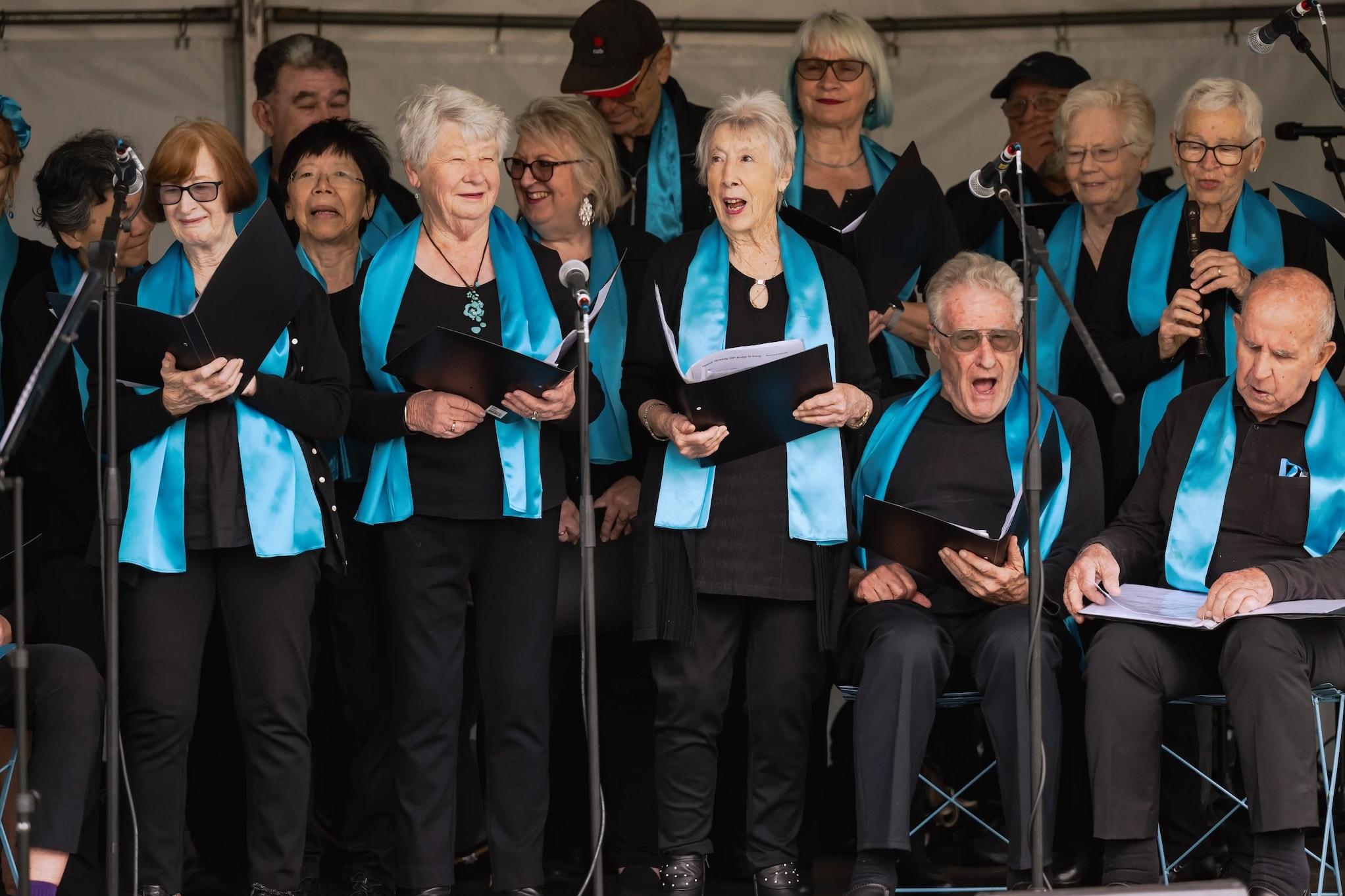 A community choir performs on an outdoor stage, with singers holding sheet music and wearing matching turquoise scarves over black outfits.