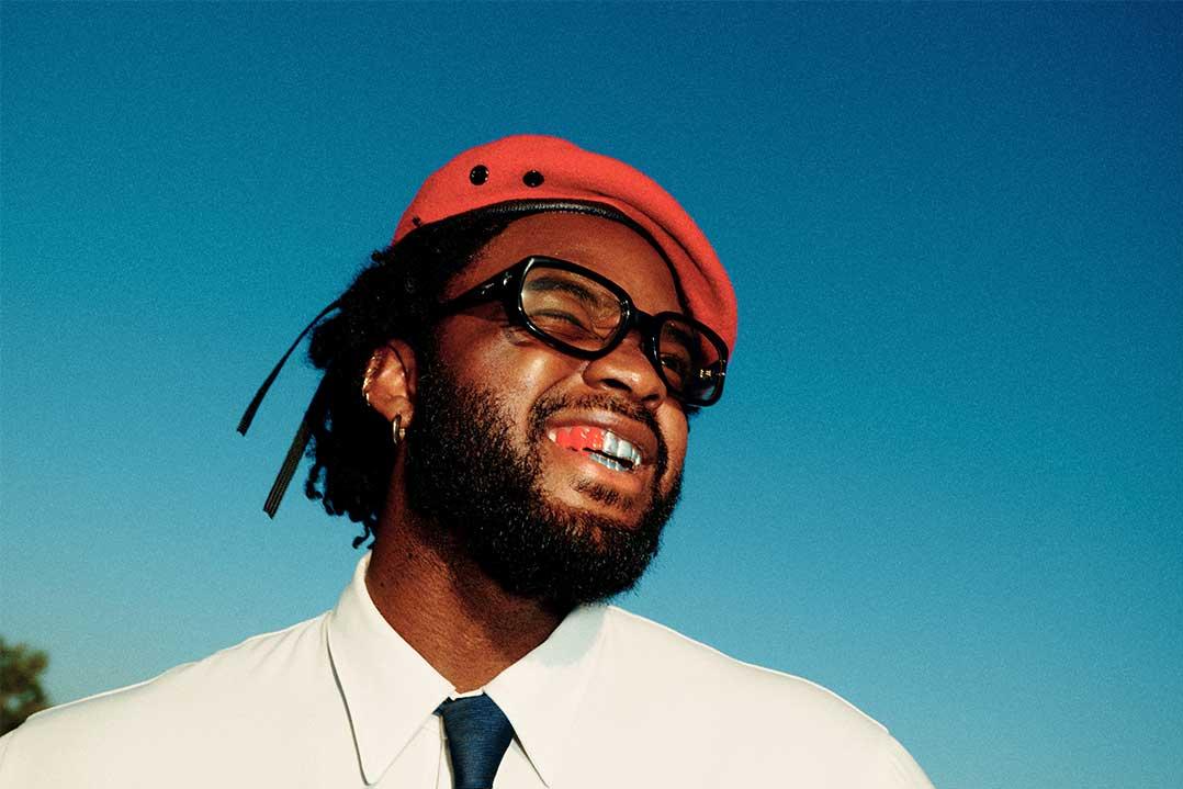 Smiling bearded Black man in a red cap and glasses, wearing a white shirt and tie against a bright blue sky; joyful, confident mood.