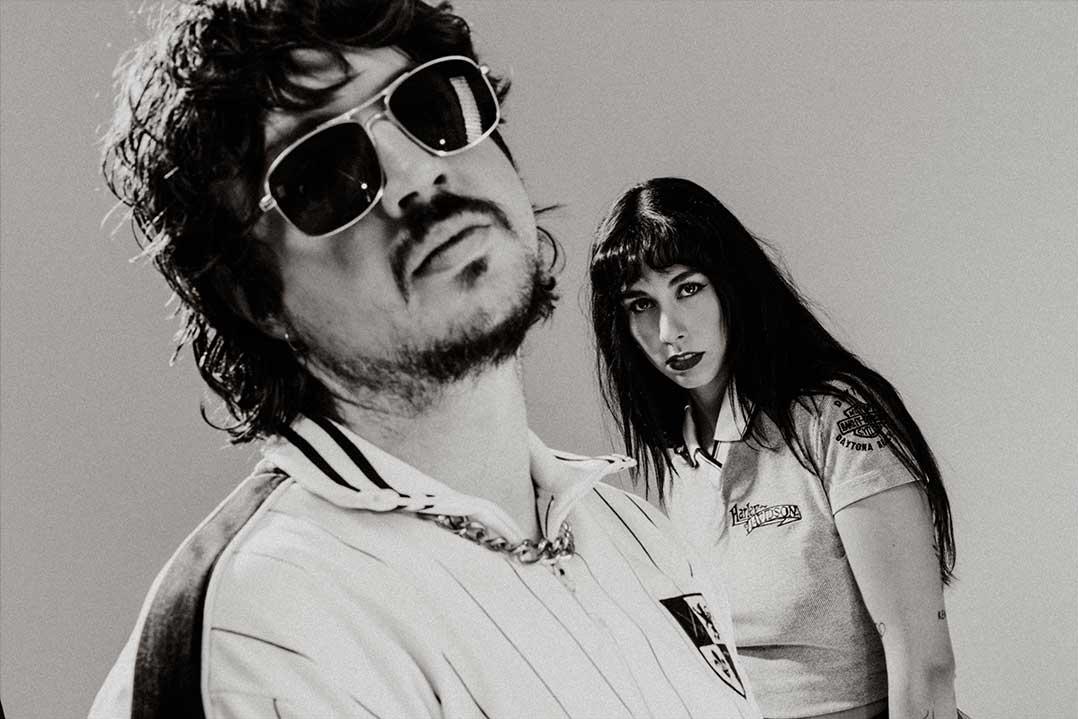 Black-and-white portrait of a mustached man in sunglasses leaning toward the camera, with a woman in a cropped polo behind him; serious mood.