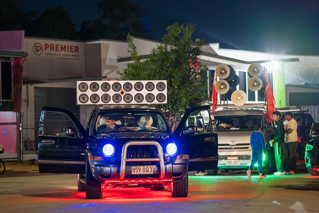 Blue vintage car with glowing headlights and roof-mounted speakers leads a vibrant nighttime car-audio gathering, with vans and people behind.
