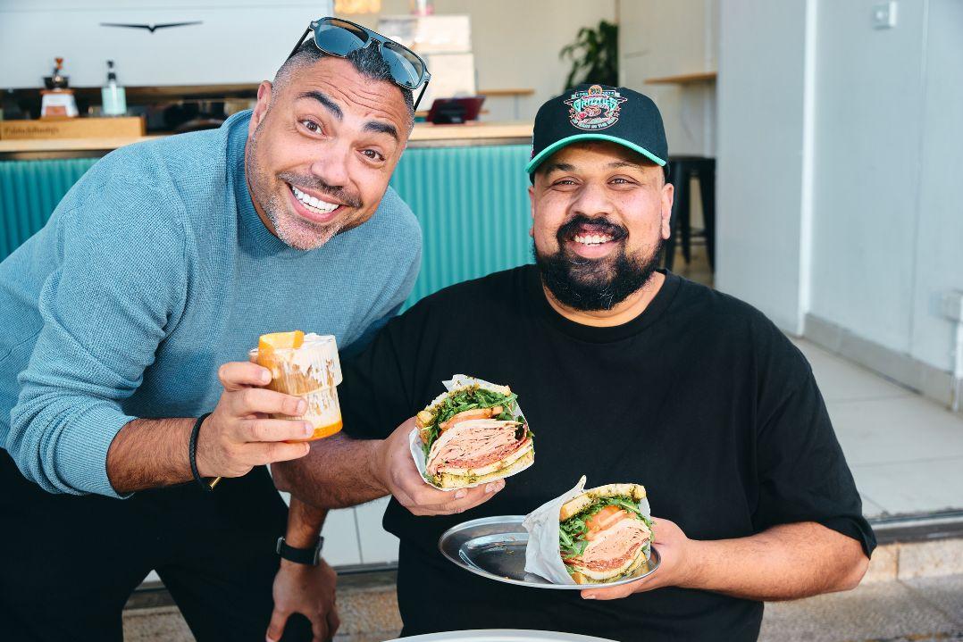 Two people seated at an outdoor café or food venue, each holding a large sandwich filled with meats, salad and condiments.