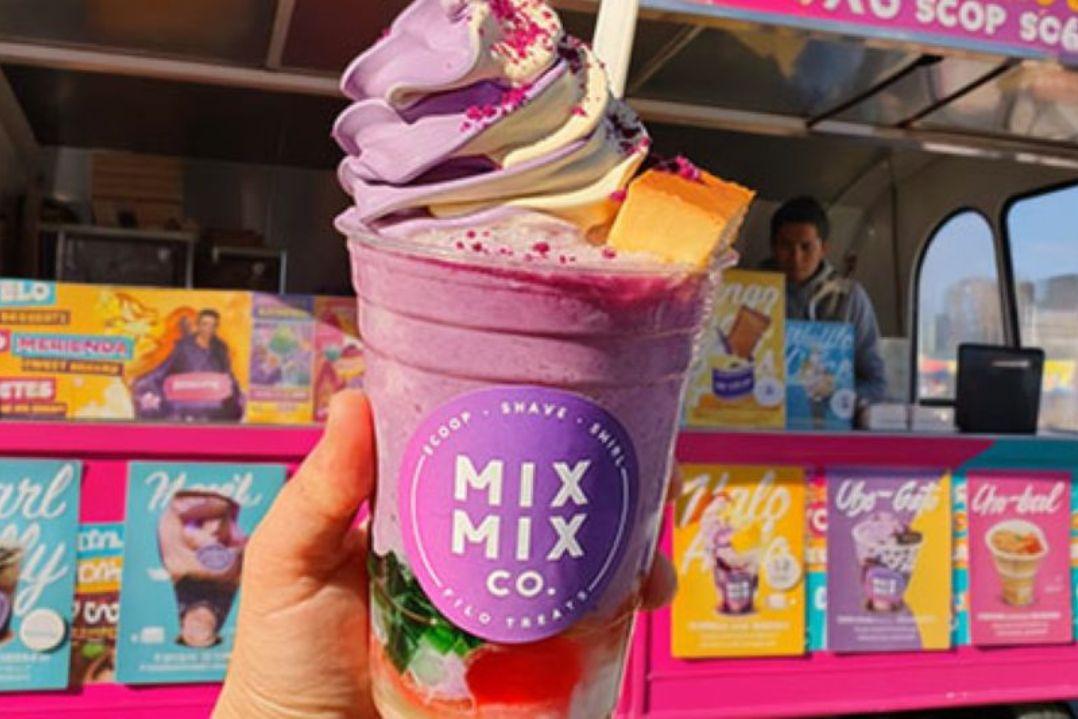 Hand holding a colourful dessert drink topped with swirled soft serve ice cream, sprinkles and cake pieces in front of a bright pink Mix Mix Co. food truck