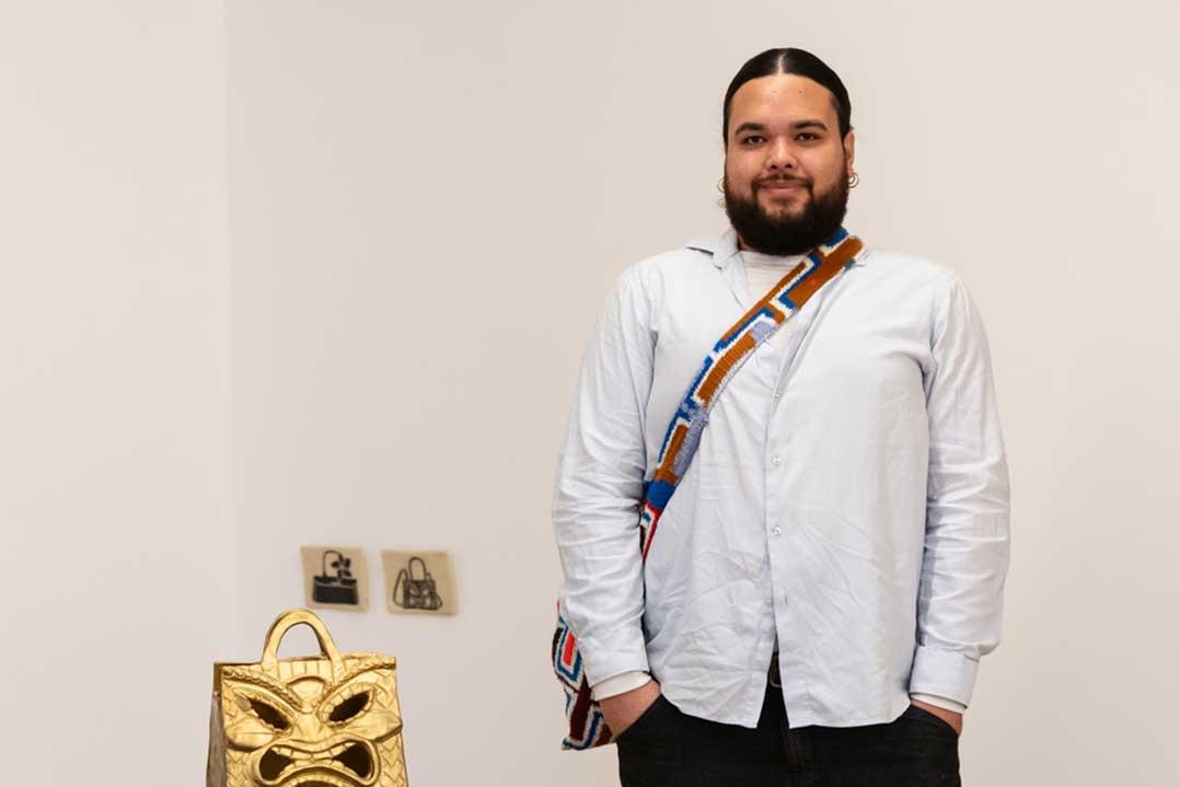 Bearded man in a white shirt and patterned crossbody bag stands beside a gold handbag sculpture in a quiet gallery, smiling calmly.